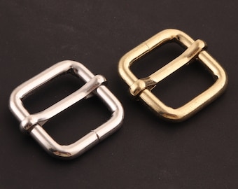 May include: Two metal belt buckles, one silver and one gold. Both buckles are square shaped with a rounded edge and a center bar.