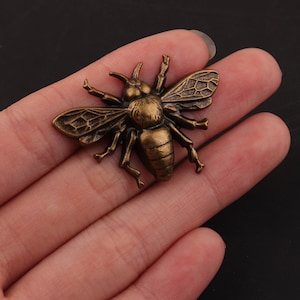 Könnte beinhalten: Eine goldfarbene Metall-Bienenbrosche mit detaillierten Flügeln und Beinen. Die Biene ist im realistischen Stil gehalten und hat eine strukturierte Oberfläche.