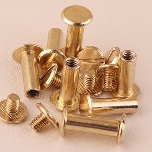 May include: A close-up of a group of brass screw-in post studs. The studs are arranged in a pile, with some of them standing upright and others lying flat. The studs are all made of a shiny, gold-colored metal.