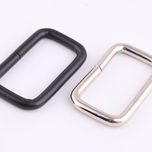 Op de afbeelding: Twee metalen D-ringen, een zwart en een zilver. De ringen zijn rechthoekig en hebben een gladde afwerking.