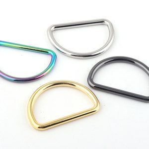 May include: Four D-rings in different metallic finishes: iridescent, silver, black, and gold. These half-circle rings are likely used for bag hardware, straps, or other crafting projects. The rings are displayed on a white background.