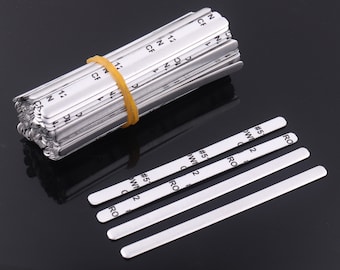 Puede incluir: Un conjunto de tiras de puente nasal de metal plateado para mascarillas, sujetas con una banda elástica amarilla. También se ven varias tiras individuales, cada una con texto impreso.