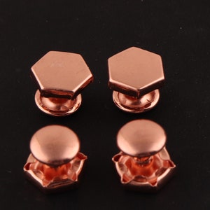 May include: Four copper-colored cufflinks. Two cufflinks have a hexagonal shape with a rounded top. The other two cufflinks have a rounded top with a hexagonal base.