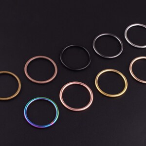 May include: Assortment of ten metal rings in various colors, including silver, gold, rose gold, and iridescent. The rings are arranged in a line on a black background.