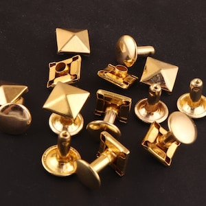 May include: A collection of gold-colored metal rivets and studs. The rivets have a flat, round head, while the studs feature a pyramid or square shape. These hardware pieces are likely for leathercraft or garment embellishment.