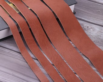 May include: Multiple strips of brown leather arranged in parallel on a wooden surface. The leather is smooth and has a subtle texture.