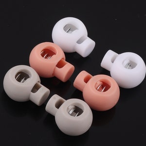 May include: Six plastic toggle stoppers in three colors: white, light brown, and light pink. Each stopper has a small metal loop for attaching to a cord or string.