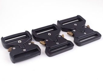 May include: Three black metal belt buckles with a brass finish. The buckles have a rectangular shape with a smooth surface and a small, rounded, brass release button.
