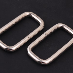 Puede incluir: Dos anillos rectangulares de metal plateado, también conocidos como anillos en D, se muestran sobre un fondo negro. Estos anillos se utilizan comúnmente para sujetar correas, cadenas u otros herrajes a bolsos, cinturones u otros accesorios.