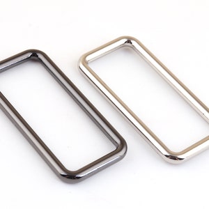 Pode incluir: Dois passadores de cinto de metal, um prateado e outro preto, com bordas arredondadas.