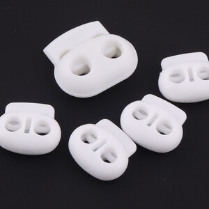 Pode incluir: Cinco travas de cordão de plástico branco, cada uma com dois furos. As travas de cordão são usadas para prender cordões ou correias.