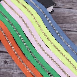 Puede incluir: Primer plano de siete cintas de cremallera de diferentes colores con dientes de metal dorado. Las cintas están dispuestas en fila, con la cinta naranja en la parte inferior y la cinta azul en la parte superior.