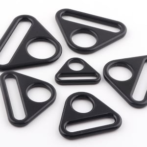 Könnte beinhalten: Sechs schwarze dreieckige Metallringe mit einem kleinen Loch in der Mitte und einer größeren ovalen Öffnung. Die Ringe sind in verschiedenen Größen.