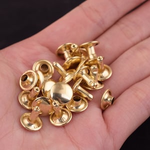 Op de afbeelding: Een close-up van een stapel gouden metalen klinknagels. De klinknagels zijn klein en rond, met een gat in het midden. Ze worden gebruikt om stof of leer aan elkaar te bevestigen.