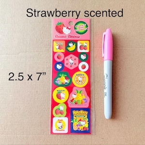 May include: A sheet of strawberry-scented stickers featuring various fruit and cartoon designs, including Hello Kitty characters. The stickers are on a red background and measure 2.5 x 7 inches. A pink and gray marker is next to the stickers.