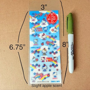 Puede incluir: Una hoja de pegatinas de Hello Kitty, de 17,1 cm de largo y 7,6 cm de ancho, con el personaje con arcoíris, flores y manzanas. Las pegatinas tienen un ligero aroma a manzana. Un rotulador Sharpie verde, de 20,3 cm de largo, está al lado de las pegatinas.