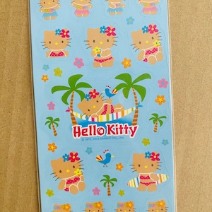 Pode incluir: Uma folha de autocolantes Hello Kitty com a personagem em várias roupas tropicais. Os autocolantes estão num fundo azul claro com palmeiras, flores e uma rede. O logótipo Hello Kitty é exibido de forma proeminente.
