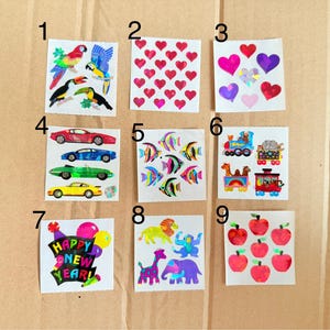 May include: A collection of nine sticker sheets featuring various designs. These include colorful birds, red hearts, multi-colored hearts, race cars, tropical fish, trains, a "Happy New Year" graphic, zoo animals, and red apples.