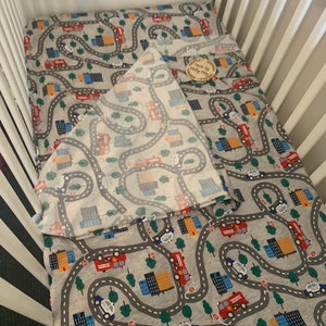 May include: A gray and white crib sheet with a city road pattern featuring fire trucks, police cars, and buildings. The sheet is folded in half and has a small wooden sign that says "Made by Molly May" on it.