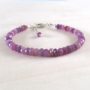 May include: A delicate bracelet featuring faceted, oval-shaped pink and purple gemstones. The bracelet is strung on a silver chain with a lobster clasp closure and a small dangling charm. The gemstones are approximately 0.25 inches in diameter.