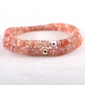 Può includere: Due braccialetti di perline color pesca-arancio. I braccialetti sono composti da piccole perline sfaccettate e sono impreziositi da una perlina argentata e una dorata. I braccialetti sono esposti su una superficie bianca.