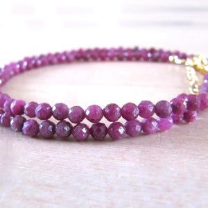 May include: A delicate necklace composed of small, faceted, deep magenta-colored gemstone beads arranged in a continuous circular strand. The necklace features a small, polished gold-toned metal lobster claw clasp closure at one end of the strand.