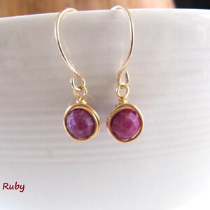 May include: Gold-coloured earrings with ruby gemstones. Each earring has a round, deep red ruby stone in a gold wire setting. The earrings hang from gold-coloured ear wires. The word "Ruby" is written in pink cursive.