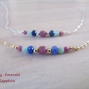 Puede incluir: Un collar de cadena dorada con una fila de piedras preciosas rubí, esmeralda y zafiro. El collar está superpuesto a una cadena de plata.