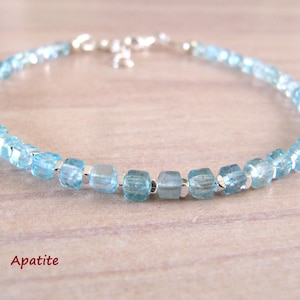 Può includere: Un delicato braccialetto con perline di apatite azzurra. Il braccialetto è infilato su una catena d'argento e ha una piccola chiusura d'argento. La parola "Apatite" è scritta in rosso in basso nell'immagine.