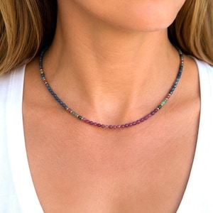 Op de afbeelding: Een delicate kralenketting met kleine, ronde, veelkleurige edelsteenkralen in tinten blauw, groen en roze, afgewisseld met kleine goudkleurige metalen tussenkralen, die in een eenvoudige, cirkelvormige streng rond de hals rust.