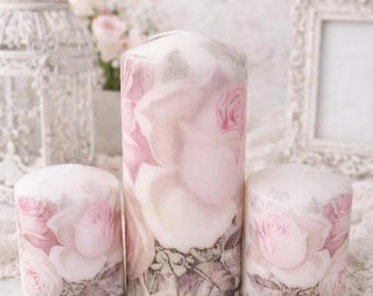 May include: Three pillar candles with a floral rose design in shades of pink and white. The candles vary in size and are arranged on a white surface with decorative elements in the background, creating a romantic and vintage aesthetic.