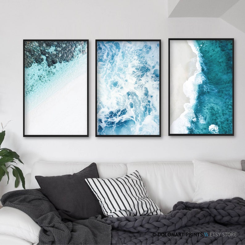 Pu&ograve; includere: Tre stampe artistiche incorniciate con viste aeree dell'oceano. Le stampe mostrano acqua turchese, spiagge di sabbia bianca e onde schiumose. Le cornici sono nere e l'opera d'arte &egrave; esposta sopra un divano bianco con cuscini e una coperta grigia.