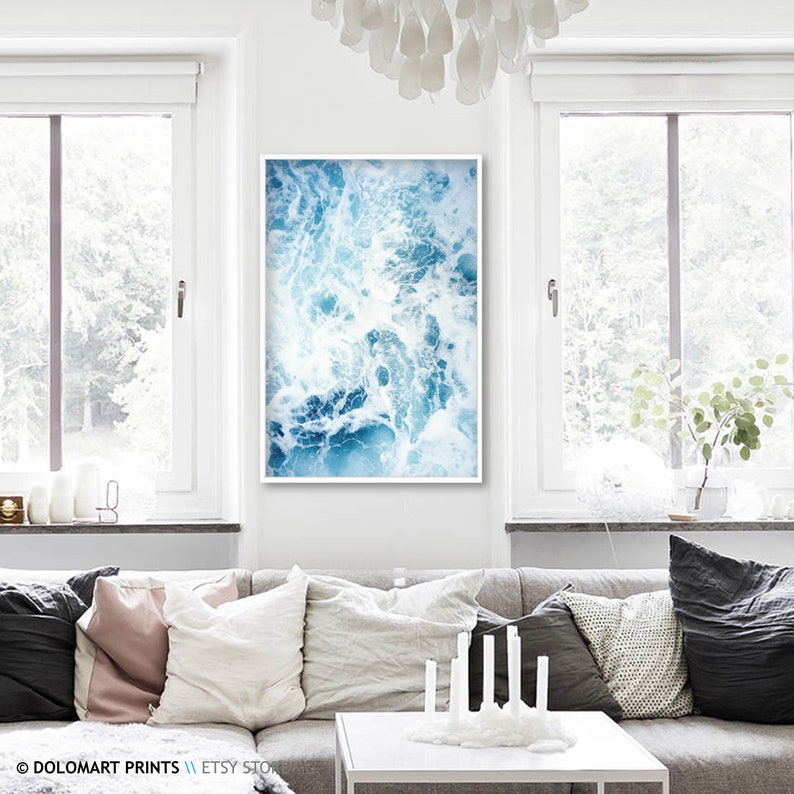 Pu&ograve; includere: Una stampa artistica incorniciata con un design astratto di onde oceaniche nei toni del blu e del bianco. L'opera d'arte &egrave; esposta in un luminoso soggiorno moderno, sopra un divano grigio con cuscini decorativi.
