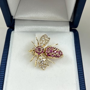 May include: A gold-toned bee brooch with ruby and diamond accents, presented in a blue jewelry box. The bee's body is composed of small, round rubies, while the wings and other details are adorned with diamonds.