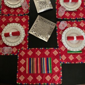 May include: Red placemats with a white knot design and a striped pattern in the center. The placemats are set on a black surface with white plates, red napkins, and clear glasses.