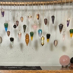 May include: A collection of pendulums hanging from a wooden shelf. The pendulums are made of various materials, including crystal, metal, and wood. The shelf is also decorated with crystals, candles, and a book titled "Light of the Wicca".