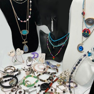 May include: A collection of necklaces, bracelets, and earrings featuring various gemstones, beads, and charms. The jewelry is displayed on mannequins and a table. Some of the pieces include a blue stone pendant, a peace sign charm, and a crescent moon charm.