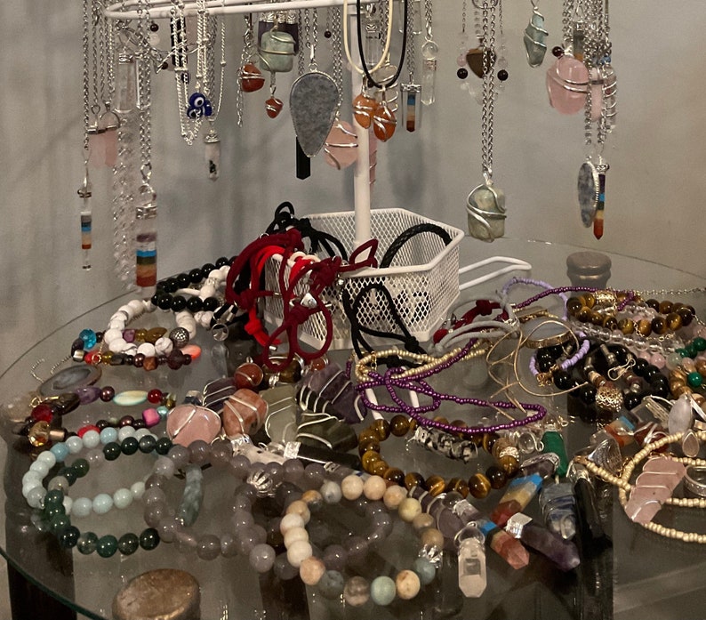 May include: A collection of colorful gemstone necklaces and bracelets, featuring various crystals, beads, and charms. The jewelry is displayed on a white stand and a glass table.