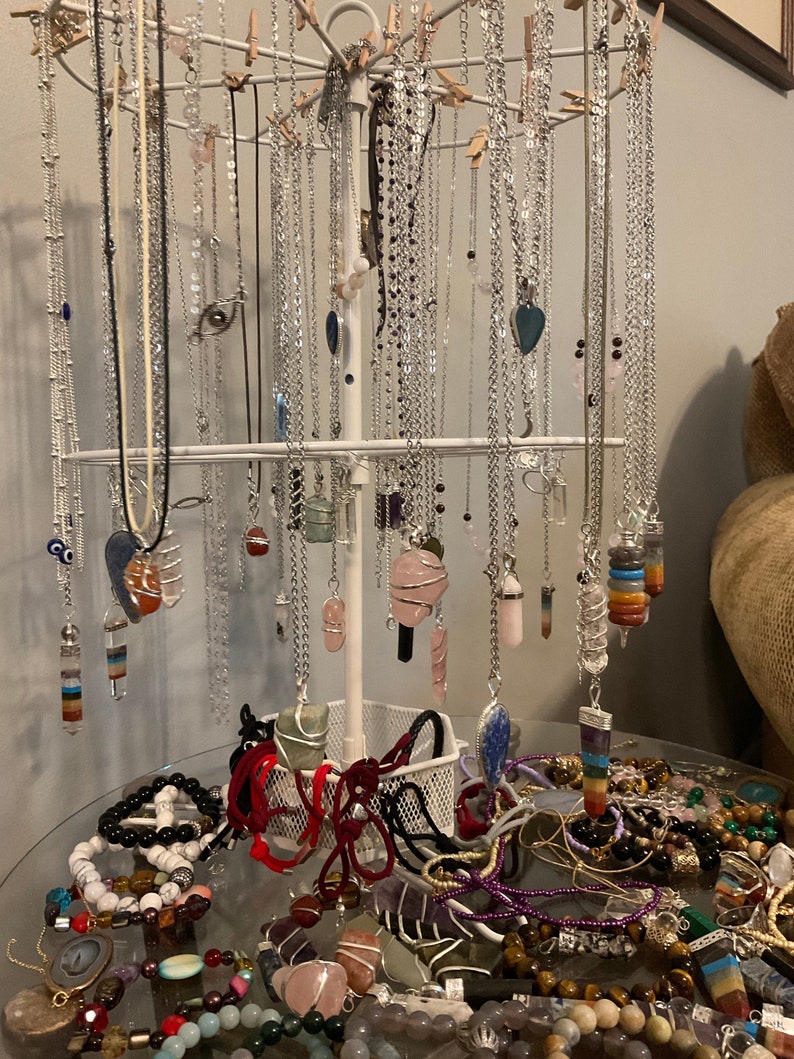 May include: A variety of crystal necklaces and bracelets on display. The necklaces are hanging on a white metal stand, while the bracelets are arranged on a glass table. The crystals are in various colors, including clear, pink, purple, blue, and green.