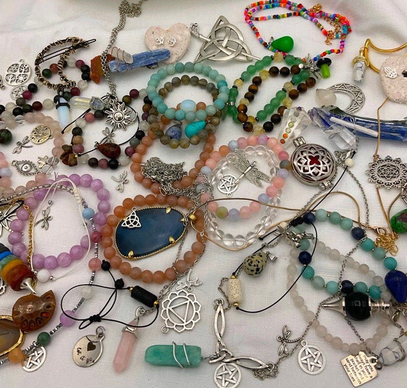 May include: A collection of colorful beaded bracelets, necklaces, and charms with various symbols and designs. The jewelry features a mix of natural stones, metal, and glass beads. Some of the charms include a dragonfly, a pentacle, and a heart.