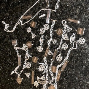 May include: A collection of silver charm necklaces and bracelets featuring pentagram, moon, and other mystical symbols. The charms are arranged on a dark background.
