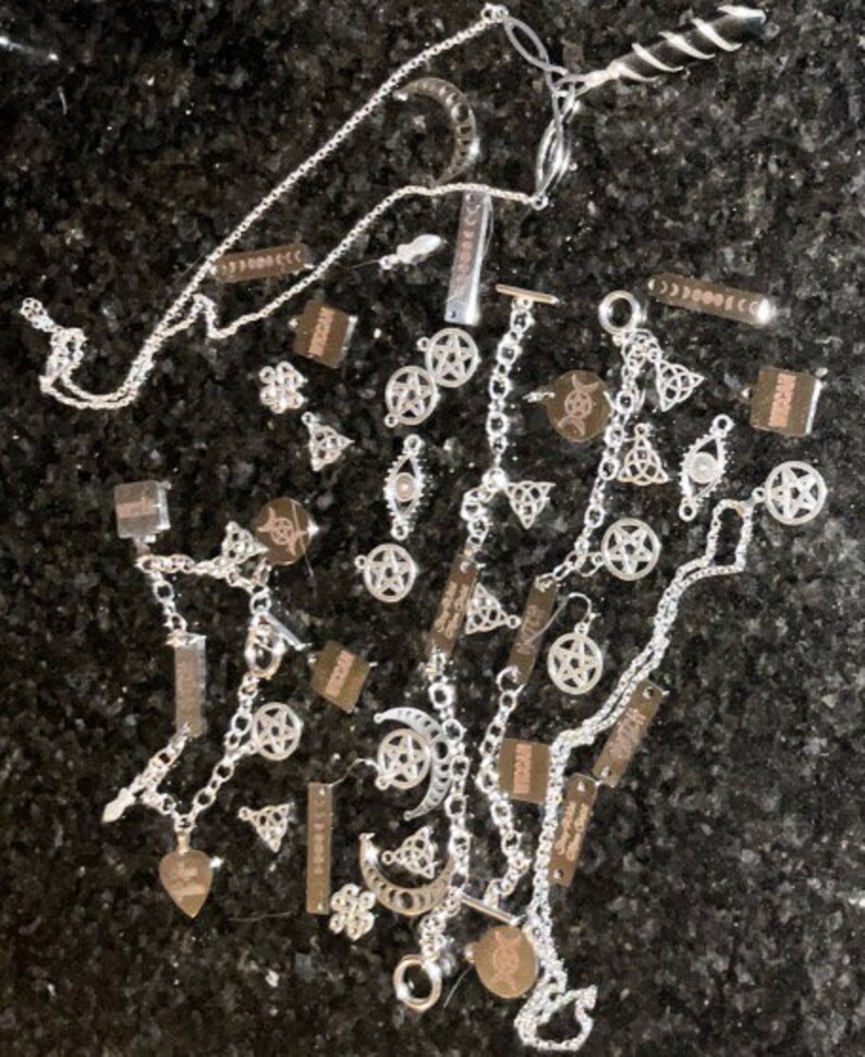 May include: A collection of silver charm necklaces and bracelets featuring pentagram, moon, and other mystical symbols. The charms are arranged on a dark background.