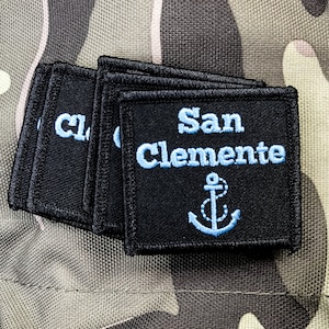 Puede incluir: Una pila de parches de tela negra con bordados azules. El parche superior dice "San Clemente" con un ancla azul debajo del texto.