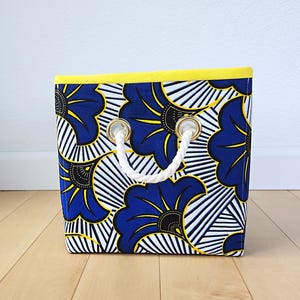 May include: A blue and yellow fabric storage bin with white rope handles. The bin has a floral pattern with black accents.