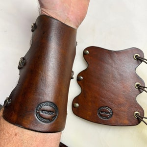 May include: A pair of brown leather arm guards with metal accents and a stamped logo that reads "Custom Leather Works".
