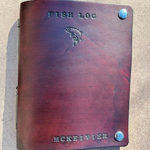 May include: A burgundy leather-bound journal with the words "FISH LOG" embossed on the front cover. A silver fish silhouette is also embossed on the cover. The journal has silver metal accents and the name "MCKEIVIER" is embossed on the bottom of the cover.