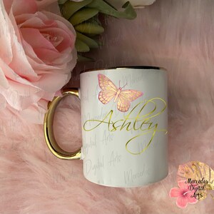 Può includere: Una tazza in ceramica bianca con bordo e manico dorati. La tazza presenta un design a farfalla rosa e il nome "Ashley" scritto in corsivo dorato.