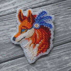 Puede incluir: Broche de zorro de cuentas con tocado de plumas azules. El zorro está representado en tonos de naranja, rojo y blanco.