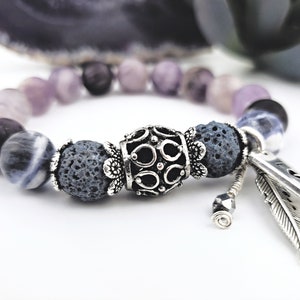 May include: A beaded bracelet featuring a mix of purple, gray, and black stones. The bracelet includes a decorative silver centerpiece and feather charms. The beads have a matte finish, and the bracelet is set against a white background.