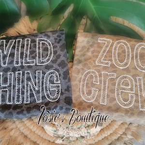 May include: Two leopard print shirts, one gray and one tan, with white stitched lettering. The gray shirt says "WILD THING" and the tan shirt says "ZOO CREW". The shirts are on a woven mat with green leaves in the background.